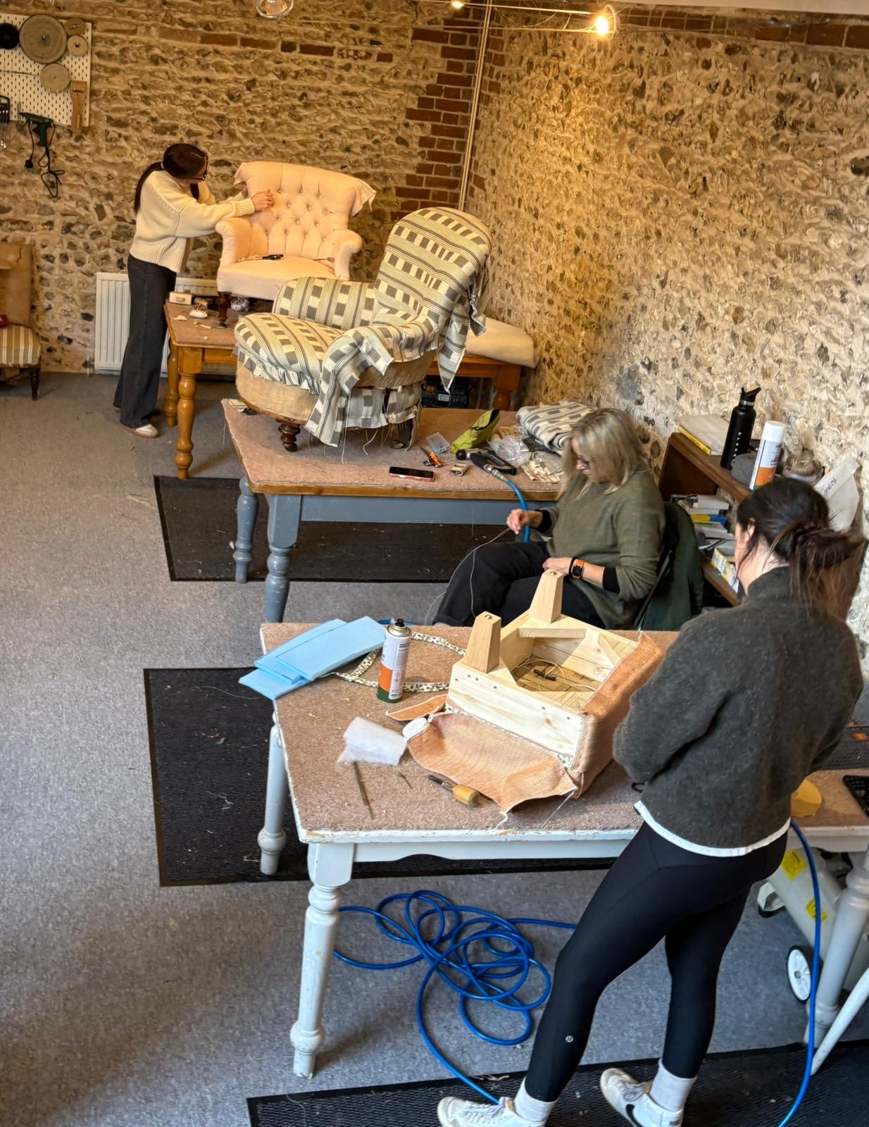 Overhead view of three students working on their upholstery projects at tables in the flint-walled studio